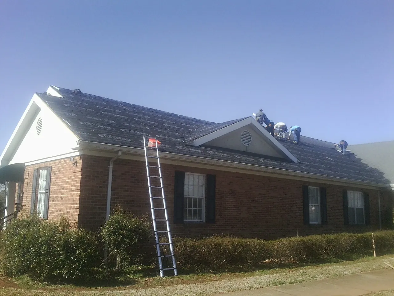Crew tearing shingles off a brick ranch roof with a ladder set — Emergency Roof Repair work by our crew in Laughlintown