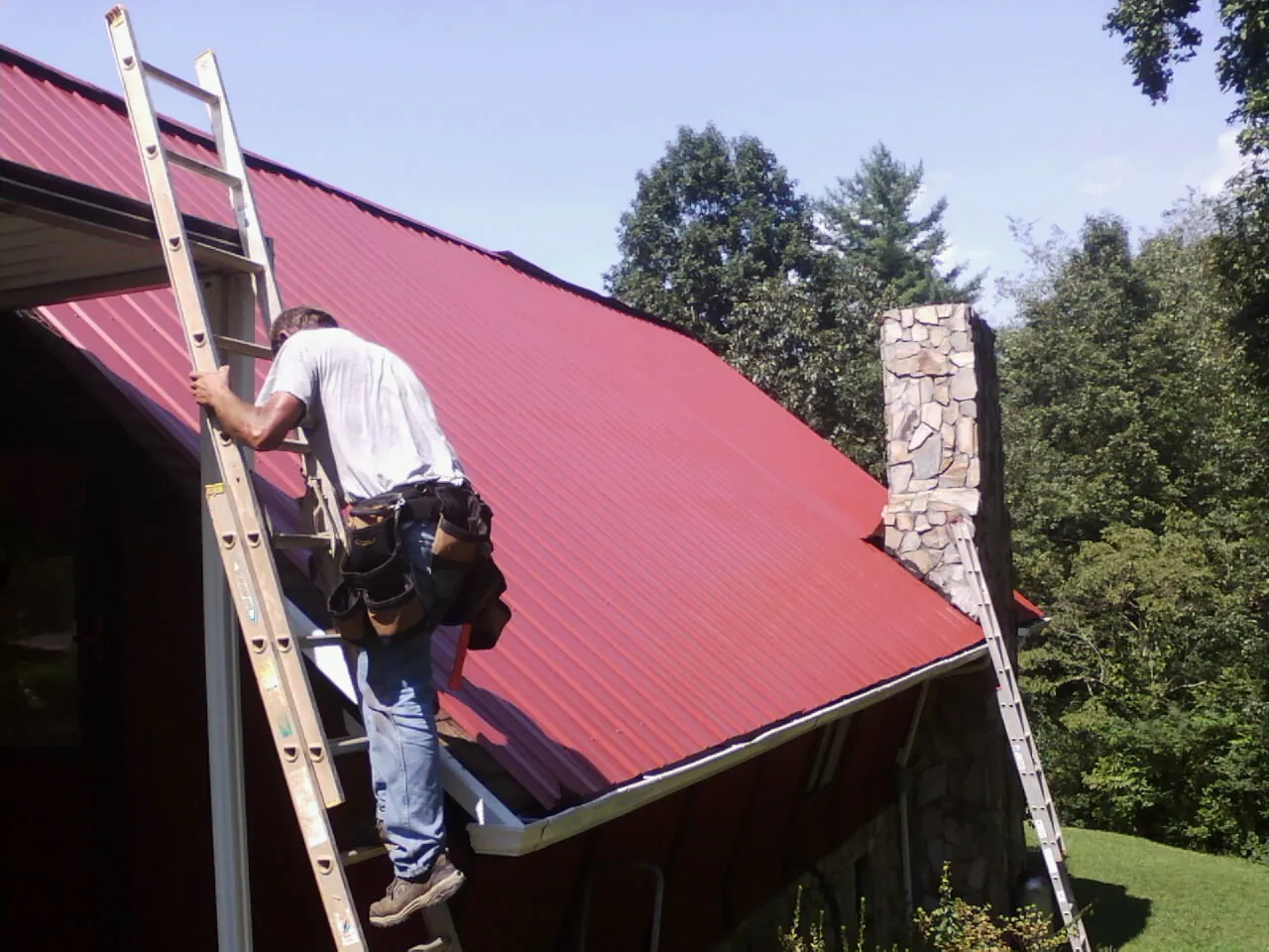 Roofer on a ladder beside a red metal roof and stone chimney — Soffit & Fascia Repair by Roofing Brothers in Laughlintown